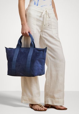 Geantă tote din suede navy cu mânere duble, textură netedă și închidere cu fermoar. Asortată cu pantaloni bej deschis într-un cadru casual.