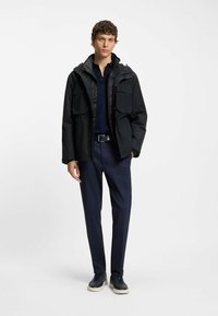 Black waterproof jacket with a hood, two front pockets, worn over a navy polo shirt and navy trousers, paired with black sneakers.