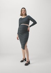 Ensemble de maternité à deux pièces rayé dans des couleurs sombres. Haut à manches longues avec taille raccourcie ; jupe ajustée avec fente sur le côté. Une paire de chaussures à talons noires.