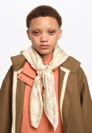 Joven con cabello corto y rizado que lleva una chaqueta marrón sobre una camiseta naranja y una bufanda crema con flores atada al cuello, mirando hacia adelante.