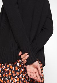 Svart stickad tröja med ribbad mudd och fåll, med en avslappnad passform. Mönstrad kjol i orange, vitt och svart synlig på den nedre halvan.