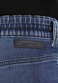 Vaqueros de mezclilla con un lavado azul oscuro, que presentan un parche de cuero con la marca "DIESEL" y detalles de costura a lo largo de la cinturilla.