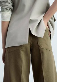 Blouse oversize gris clair avec un détail de dos fendu, assortie à un pantalon à jambes larges vert olive avec des poches arrière.