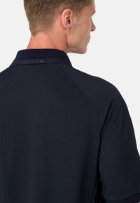 Jūras spēku polo krekls ar teksturētu apkakli un smalku logo aizmugurē, kuram ir gluda auduma struktūra un raglānu piedurknes mūsdienīgai piegrieztei.