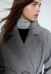 Grauer Wollmantel mit Reverskragen, tailliertem Gürtel und strukturiertem Stoff; darüber ein grauer Rollkragenpullover und ein schwarzes Oberteil getragen.