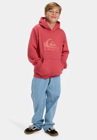 Sudadera con capucha roja de algodón con logotipo frontal, bolsillo delantero y puños acanalados. Combinada con vaqueros holgados azul claro y zapatillas negras.