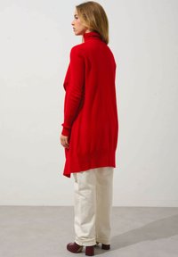 Rote Strickjacke mit hohem Kragen, ausgestattet mit gerippten Bündchen und Saum. Kombiniert mit off-white weiten Hosen und bordeauxfarbenen Absatzschuhen.