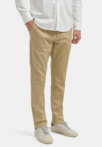 Beige bomullbyxor med skräddarsydd passform, sidofickor och minimalistiska sömdetaljer, tillsammans med ljusgråa sportskor.