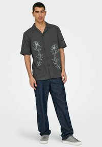 Korte mouwen overhemd van donkergrijze stof, met lichte blauwe bloemenborduurwerk op de voorkant, gecombineerd met loszittende denim broek.