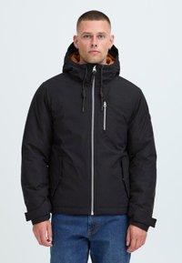 Man wearing a black hooded winter jacket with a front zipper and blue jeans, standing against a plain light background.