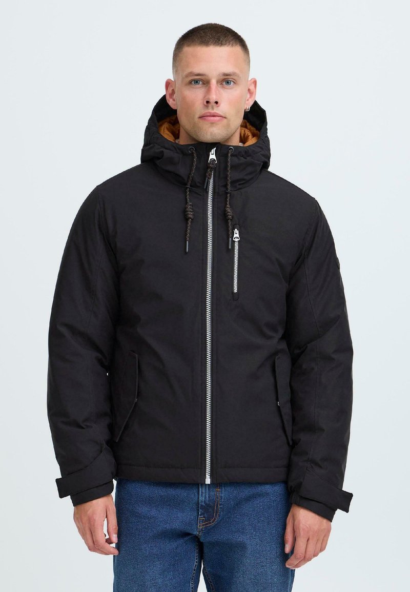 Man wearing a black hooded winter jacket with a front zipper and blue jeans, standing against a plain light background.