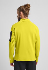 Maglietta sportiva a maniche lunghe di un brillante giallo, in tessuto texturizzato, con colletto alto e un accento grigio scuro sulle spalle.