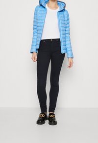 Giacca imbottita azzurro chiaro con cappuccio, t-shirt bianca, jeans skinny scuri e scarpe nere con dettagli in catena dorata.