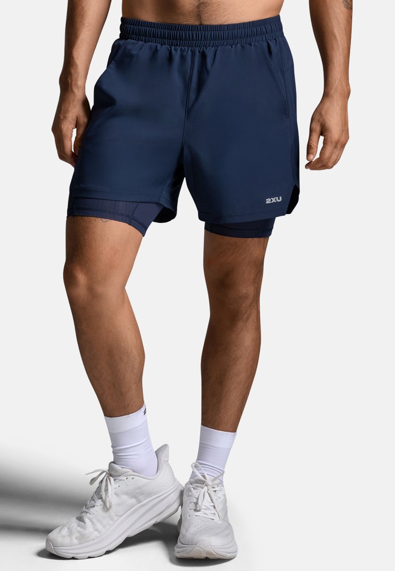 Granatowe sportowe spodenki o lekkiej, gładkiej fakturze. Charakteryzują się średnią długością i siateczkową podszewką, zestawione z białymi butami do biegania.