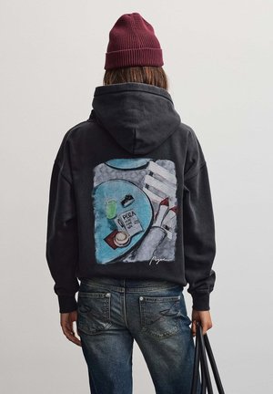 Persona che indossa una felpa nera con cappuccio con una grafica colorata di un tavolo, caffè e gambe incrociate sulla schiena, abbinata a jeans blu e un berretto bordeaux.