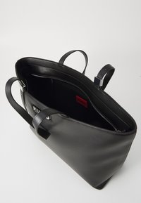 Black leather tote bag with dual handles, zippered compartment, and a red "HUGO" label inside, featuring a smooth texture and minimal design.