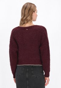 Burgundowy sweter dzianinowy o teksturowym wykończeniu, z szerokimi rękawami i okrągłym dekoltem. Posiada srebrny akcent na dolnym brzegu z tyłu.