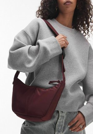 Kvinde iført grå oversized sweatshirt og jeans, der holder en bordeaux skuldertaske med lynlåslukning.