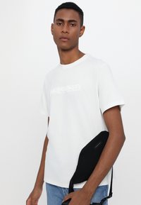 White cotton T-shirt with embossed "MICHAEL KORS" logo. Paired with blue jeans and a black shoulder bag featuring a label.