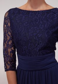 Robe bleu marine avec corsage en dentelle et manches trois-quarts, dotée d'une taille plissée et d'une jupe fluide. Motif floral en dentelle complexe.