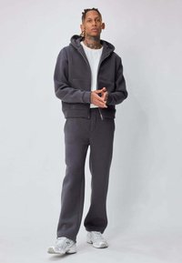 Sweat à capuche gris à fermeture éclair avec une poche avant, assorti à un pantalon. T-shirt blanc en dessous. Modèle portant des chaussures de sport gris clair.