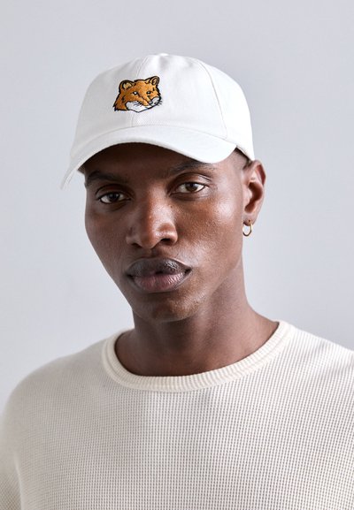 Casquette de baseball blanche avec un design de tête de renard brodé. La casquette a une visière incurvée et est assortie à un pull beige texturé.