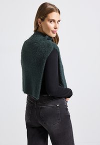 Groene fuzzy vest gemaakt van zacht materiaal, gedragen over een zwart lang mouwen shirt. Het model combineert het met donkergrijze jeans.