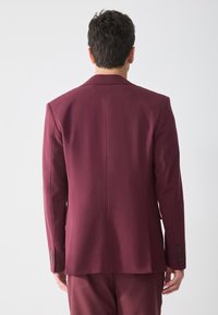 Chaqueta de traje burdeos con solapa muesca, corte ajustado, tela suave y detalle de puño con cuatro botones. Vista trasera, sin patrones visibles.