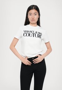 Balti medvilnės marškinėliai su užrašu "VERSACE JEANS COUTURE" ryškiomis juodomis raidėmis. Modelis vilki juodas džinsus, rankas laikydamas ant klubų.