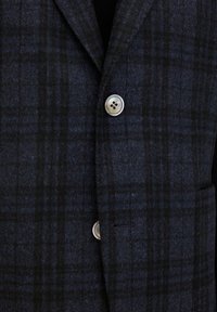 Mörk rutig kavaj gjord av texturerad ull, med notched krage, silverknappar och en framficka. Färgerna inkluderar mörkblå och svart.