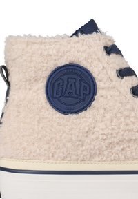 Béžové vysoké tenisky s huňatou texturou, námořnické šňůrky a kulatá námořnická nášivka s nápisy "GAP" a "1969."
