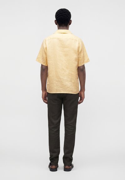 Homme debout de dos, portant une chemise jaune pâle à manches courtes, un pantalon foncé et des sandales noires sur un fond clair uni.