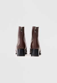 Stivali alla caviglia in pelle marrone con tacco a blocco, dotati di una chiusura con zip sul retro e una superficie liscia e elegante. Base del tacco nera piatta.