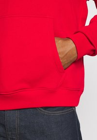 Rotes Sweatshirt mit Kängurutasche, gerippten Bündchen und glatter Textur, kombiniert mit dunklen Jeans aus Denim.