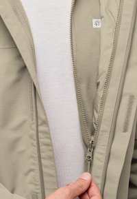 Giacca beige con zip, caratterizzata da uno strato esterno testurizzato, morbido interno in pile e fodera in tessuto a contrasto. La cerniera e il tirante sono visibili.