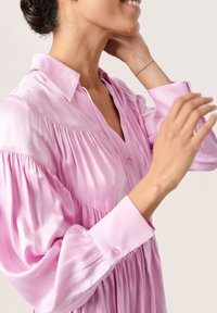 Blusa in satin rosa con colletto abbottonato, maniche arricciate e pieghe verticali sul davanti, caratterizzata da una vestibilità comoda e una superficie liscia.