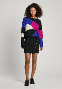 Strickpullover mit lila, rosa, weißen und schwarzen geometrischen Mustern; strukturierter Stoff. Schwarze Minirock mit verstreuten metallischen Akzenten. Schwarze Slipper.