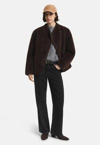 Braune Fleecejacke mit rundem Kragen, getragen über einem grauen Strickpullover. Kombiniert mit schwarzen weit geschnittenen Jeans und braunen Ankle Boots. Hellbraune Mütze.