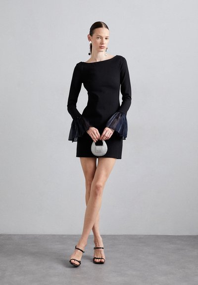 Black long-sleeve dress with sheer bell sleeves, bodycon fit, and above-knee length. Accessorized with a silver round handbag and black heels.