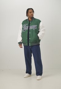 Casaco varsity verde e branco com "Tommy" em letra cursiva, apresentando botões de pressão, riscas nas mangas, usado com jeans azuis e ténis brancos.