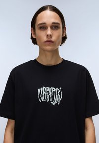 Svart bomullst-skjorte med et fremtredende, stilisert, hvitt logo på forsiden. Klassisk rund hals og korte ermer.