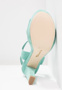 Sandale à talons hauts en daim vert clair avec semelle beige, nom de la marque Tamaris imprimé sur la semelle, vue de dessous.