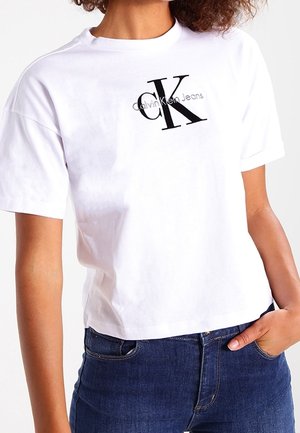 Persoon die een wit Calvin Klein Jeans T-shirt met zwart logo en blauwe spijkerbroek draagt, één hand in de zak, tegen een effen achtergrond.