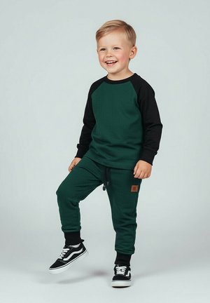Uśmiechnięty młody chłopiec stojący z jedną uniesioną stopą, ubrany w zielono-czarną bluzę z długim rękawem, pasujące spodnie oraz czarne trampki z białymi podeszwami.
