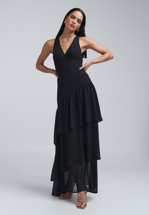 Black halter maxi dress with layered ruffles, v-neckline, and a flowy silhouette; features a smooth texture and subtle sheen. Gold strappy sandals.