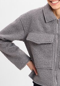 Szara polarowa kurtka z zapięciem na zamek, kołnierzykiem typu point oraz dwoma naszytymi kieszeniami. Tekstura jest miękka, z przytulnym, puszystym wyglądem.