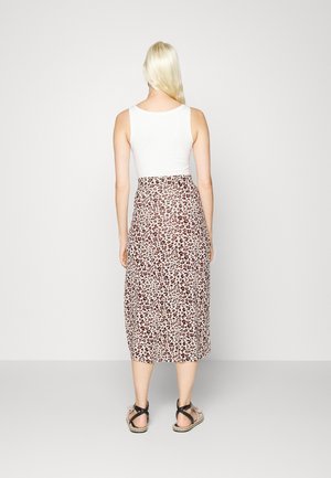 Lyshåret kvinde står med ryggen mod kameraet iført hvid ærmeløs top, leopardprint midi-nederdel og sorte sandaler med stropper på gråt gulv.
