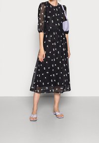 Schwarzes Midi-Kleid mit floralem Stickerei, durchsichtigen Puffärmeln und geraffter Taille. Getragen mit helllila flachen Sandalen und einer passenden Tasche.