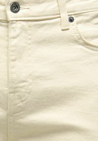 Gros plan sur un pantalon en denim beige montrant un bouton métallique, un rivet, une boucle de ceinture et les détails des coutures de la poche avant.