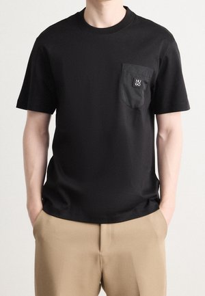 Personne portant un T-shirt noir à manches courtes avec une poche poitrine gauche étiquetée "HUGO" et un pantalon beige, les mains dans les poches.